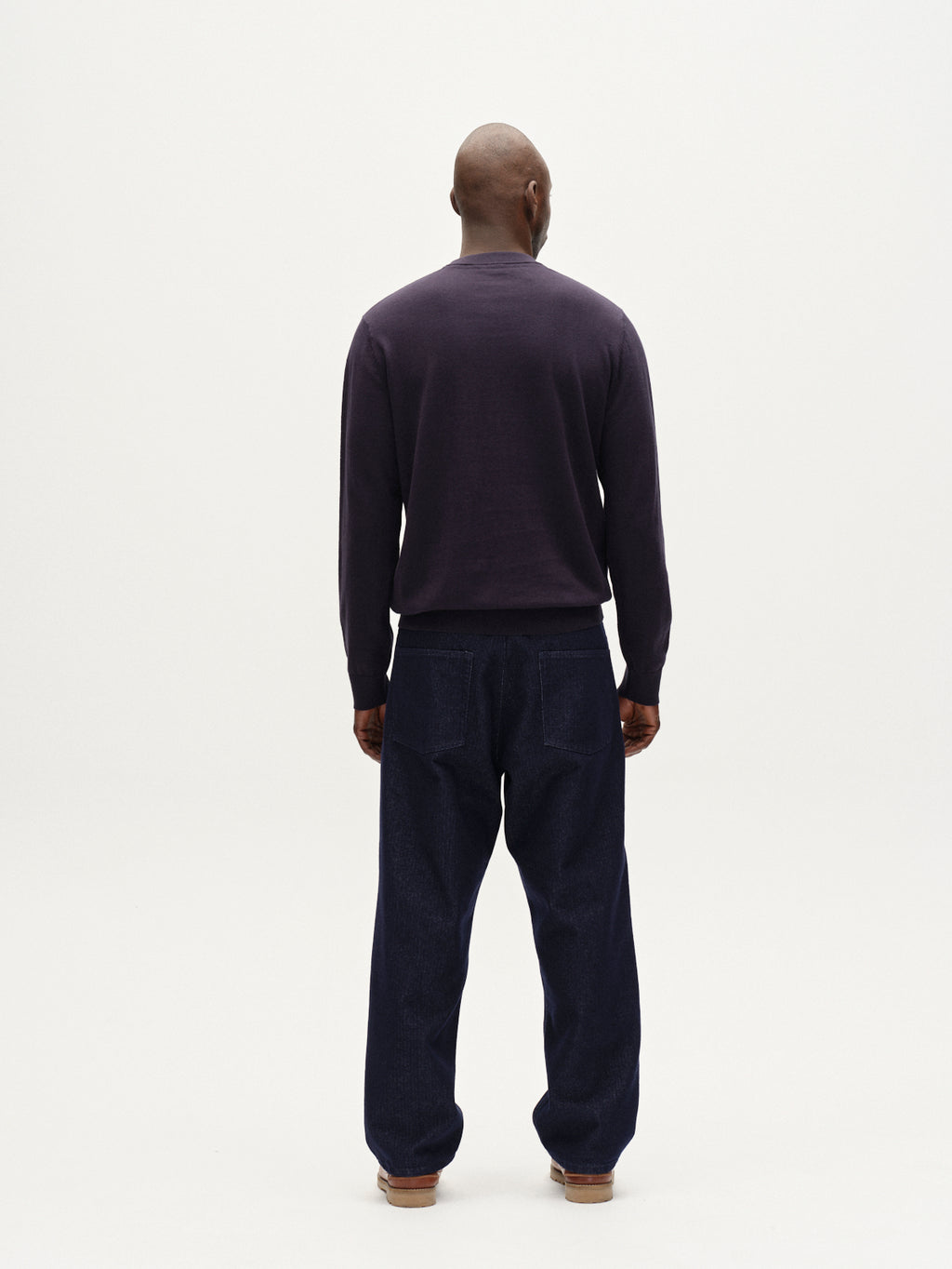 A person faces away from the camera wearing the Gabba Amori Prex LS - Navy sweater and mens regular fit, loose dark blue jeans against a plain white background.
