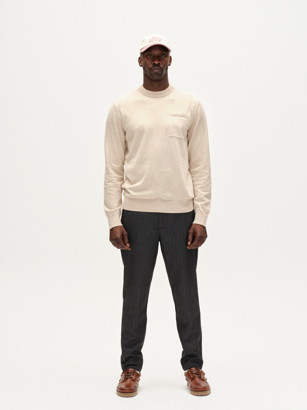 A man stands against a white background wearing Gabbas Amori Prex LS - Sand sweater, dark gray pants, brown shoes, and a white baseball cap. He faces forward with a neutral expression and relaxed posture.