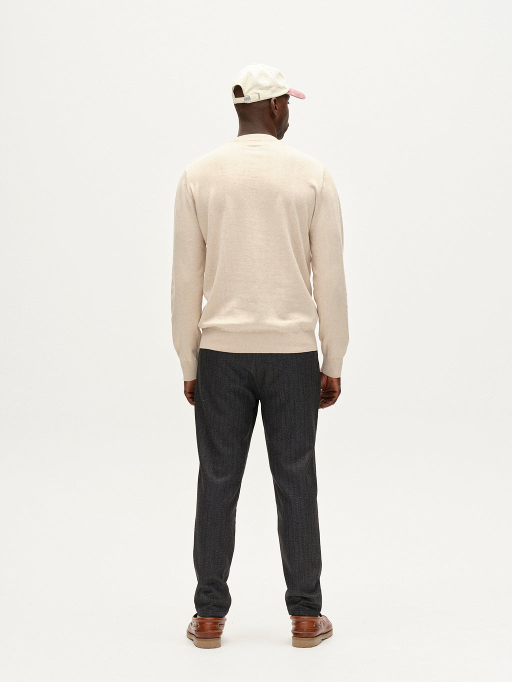 A man stands facing away from the camera against a plain white background, wearing the Gabba Amori Prex LS sweater in sand, dark gray pants, brown shoes, and a white baseball cap with a pink brim.