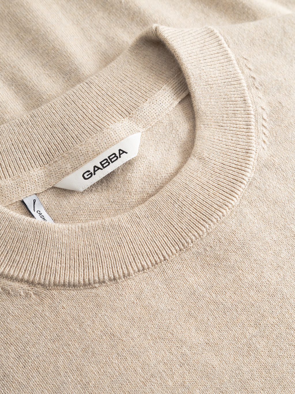 A close-up of the Amori Prex LS - Sand sweater shows its beige knit crew neck and ribbed cuffs. Inside the collar, a white label features the Gabba brand name in black letters.