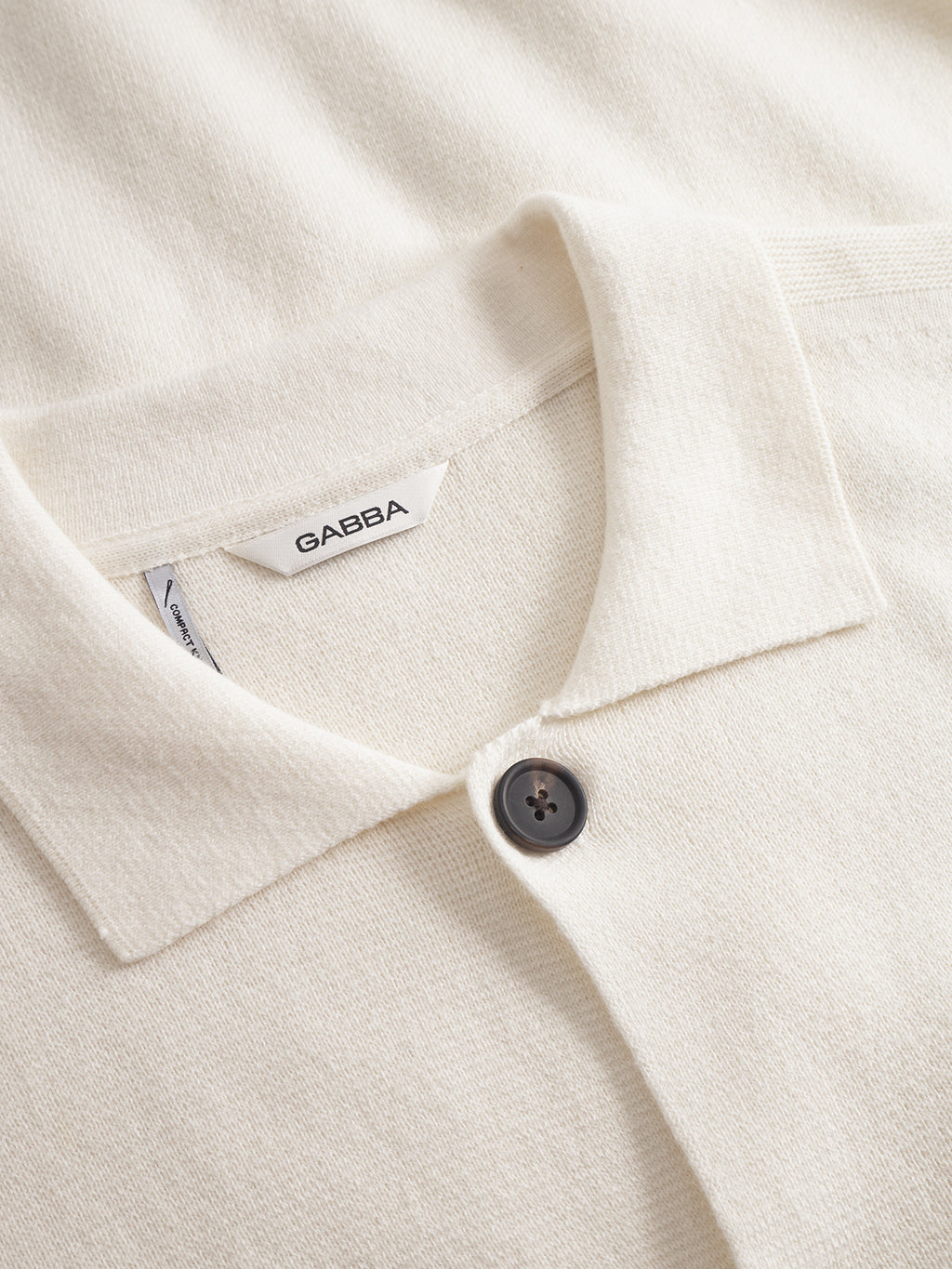 Close-up of the Gabba Blanko New Cardi LS in off white, crafted from soft, textured fabric with a single dark button and a white GABBA label—a minimalist design ideal for layering or wearing solo.