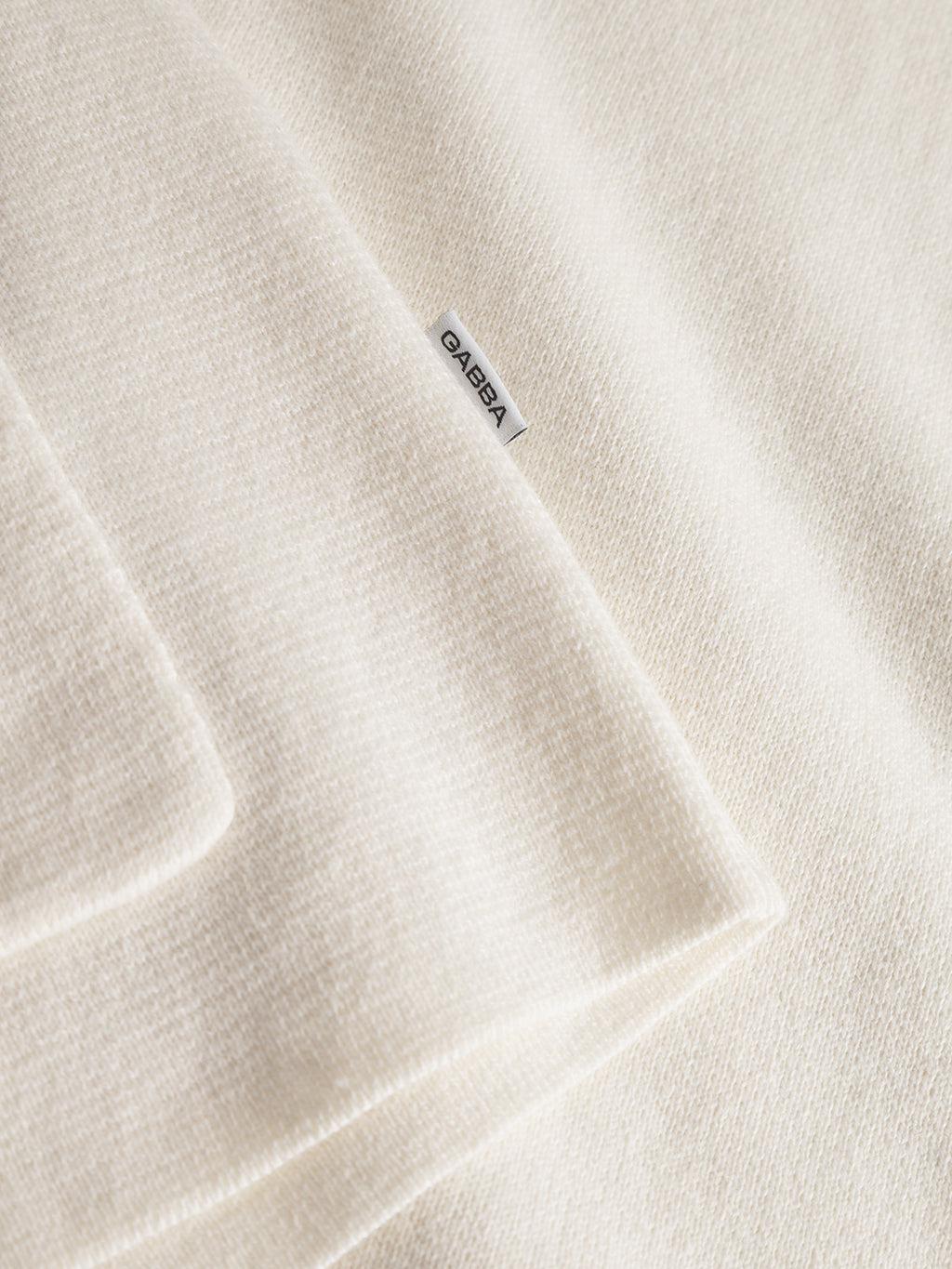Close-up of the Gabba Blanko New Cardi LS in off white, featuring a soft texture and minimalist design. A small white Gabba label is sewn on the edge, making this cream-colored cardigan perfect for effortless layering.
