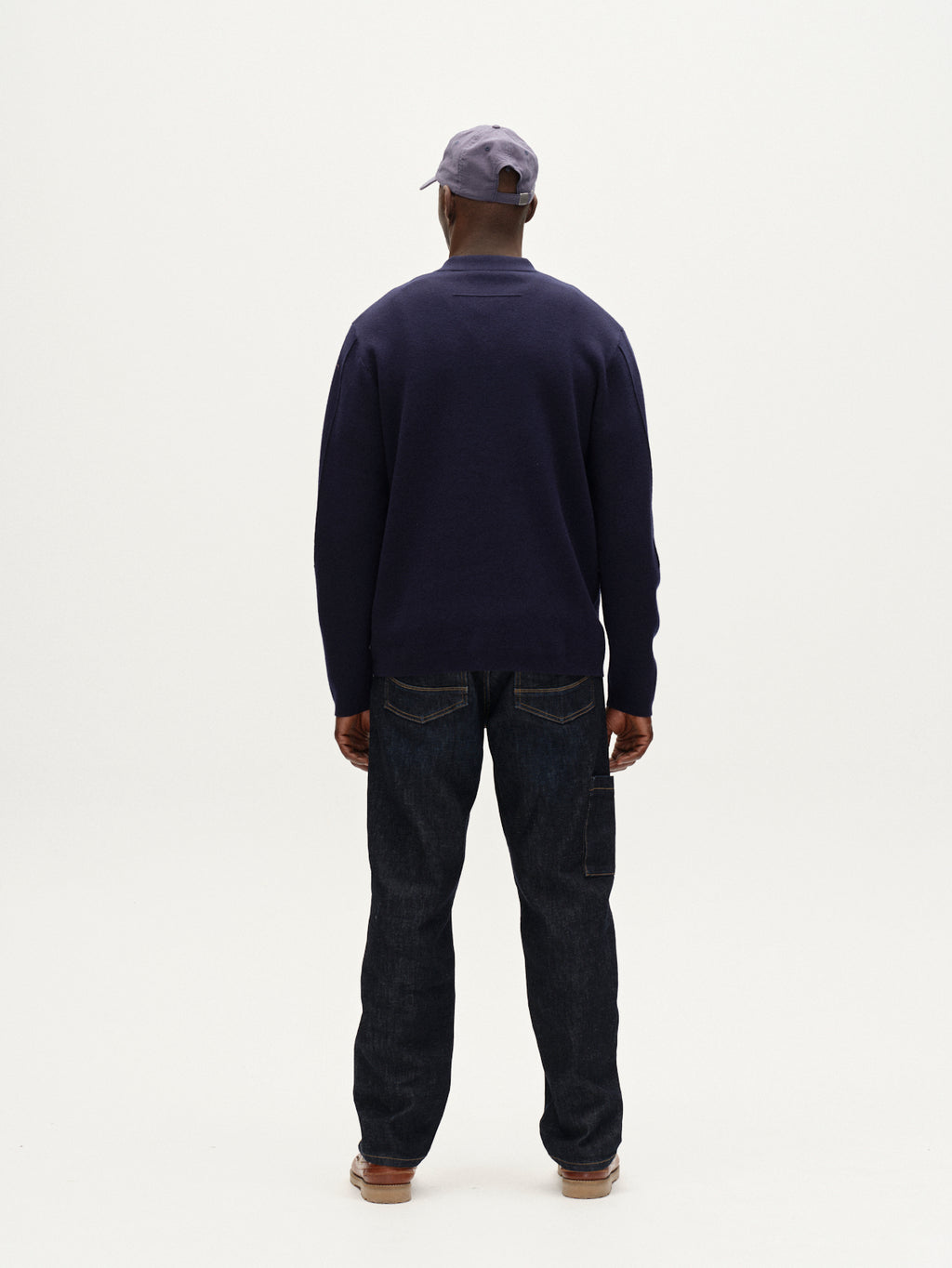 Person wearing a navy sweater and dark jeans with a cap on a white background