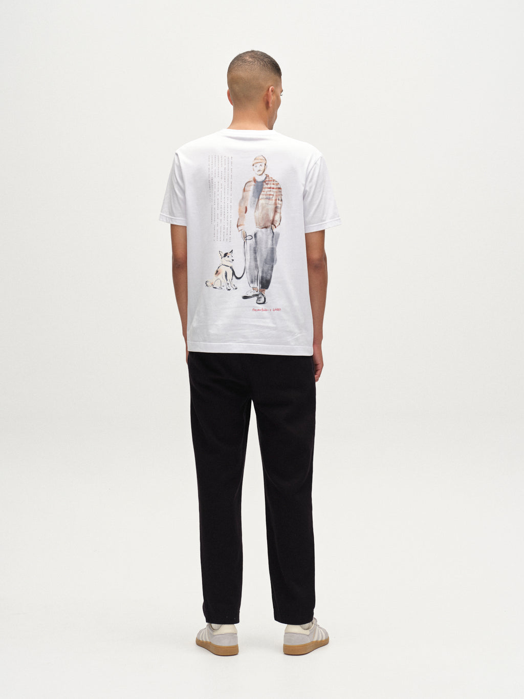 A person faces away wearing the Gabba Dune Kay Man Bauw SS GOTS t-shirt in white organic cotton, featuring an illustrated man and dog on the back, paired with black pants and beige sneakers against a plain white background.