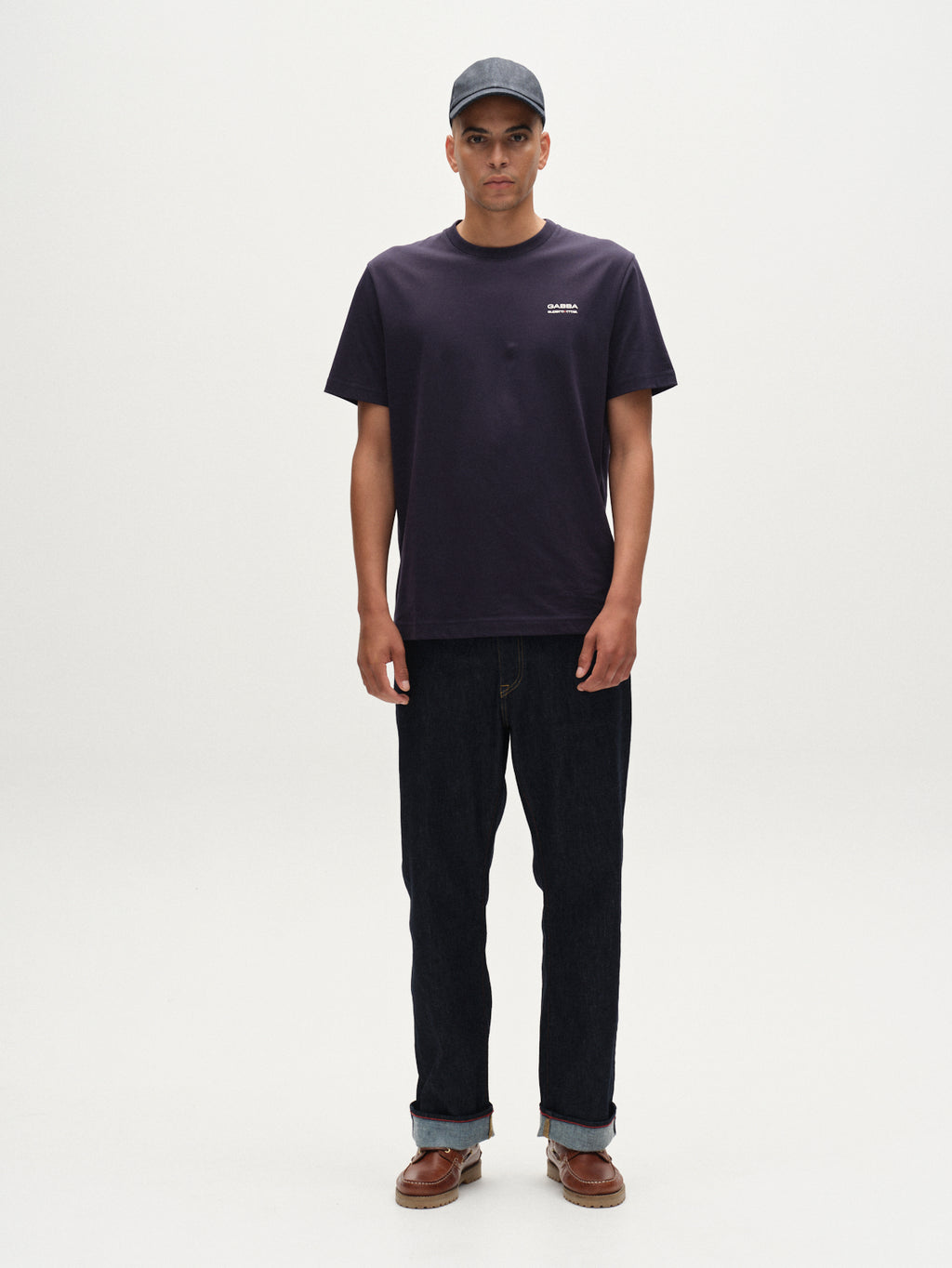 A man stands facing forward against a plain white background, wearing the Gabba Dune Kay Man Bauw SS GOTS T-shirt in navy, a black baseball cap, dark jeans, and brown shoes.