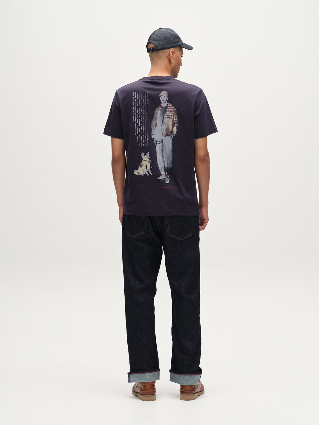 A man in a dark cap, dark jeans, brown shoes, and Gabba’s Dune Kay Man Bauw SS GOTS T-shirt in navy—with a graphic of a man and dog on the back—stands facing away in a minimalist white studio.