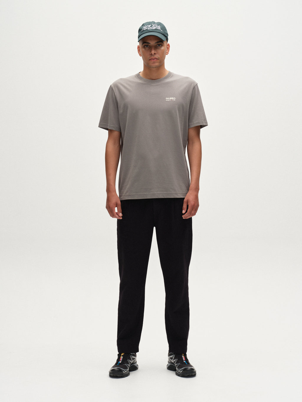 A person stands facing forward against a white background, wearing a Gabba Dune Kay Man Bauw SS GOTS T-shirt in Dust Sage Green, along with black pants, black sneakers, and a dark baseball cap.