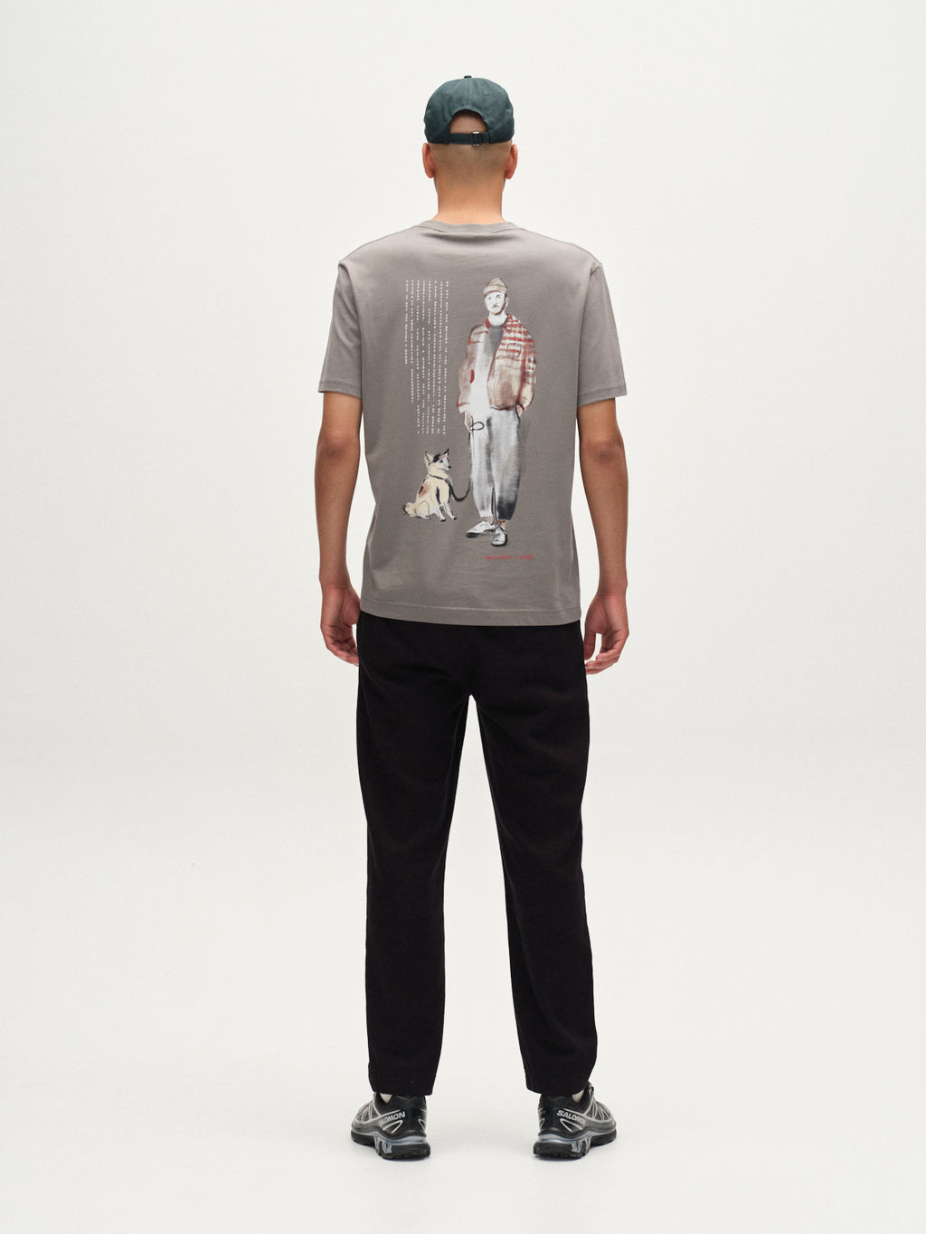 A person faces away, wearing a Gabba Dune Kay Man Bauw SS GOTS T-shirt in Dust Sage Green made from organic cotton, paired with black pants and sneakers. The shirt features a man and dog illustration with text, set on a white background.
