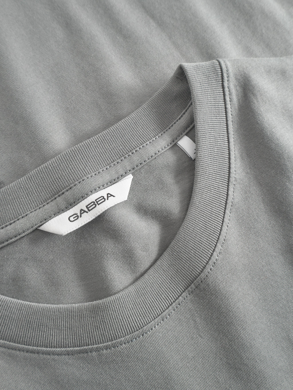 Close-up of the Dune Kay Man Bauw SS GOTS T-shirt by Gabba in Dust Sage Green. Features a visible collar, white Gabba label inside the neckline, and is crafted from smooth, soft GOTS-certified organic cotton with gentle folds.