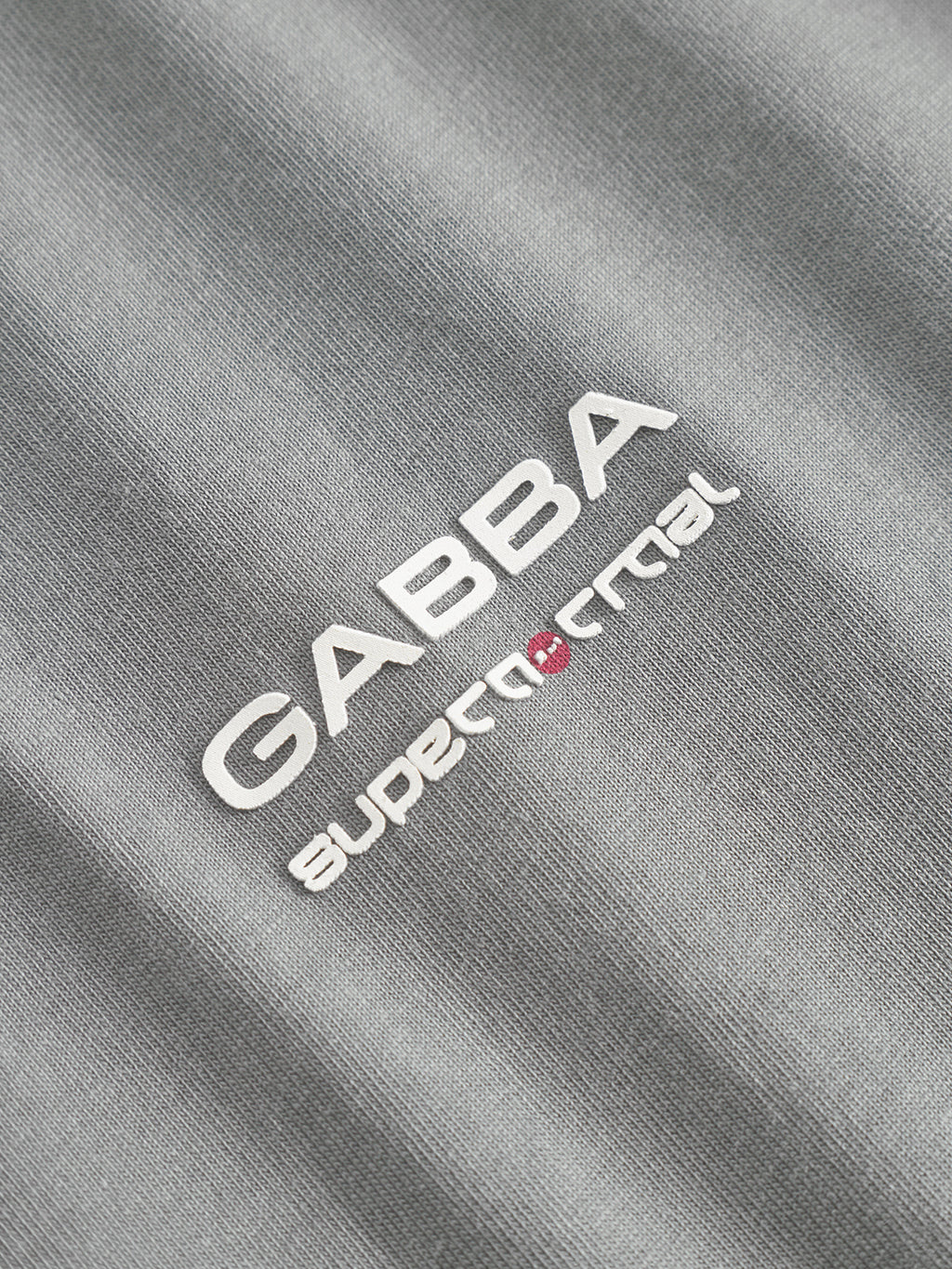 Close-up of Gabba’s Dune Kay Man Bauw SS GOTS tee in Dust Sage Green, featuring white “GABBA” above “supernatural” text, with the a in supernatural replaced by a small red design. Made from GOTS-certified organic cotton.