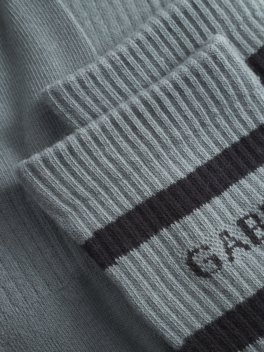 Close-up of a dust sage green ribbed knit fabric from Gabba’s Zino Socks, featuring two black horizontal stripes and partial GAB text. The cotton blend texture and stitching details are clearly visible.