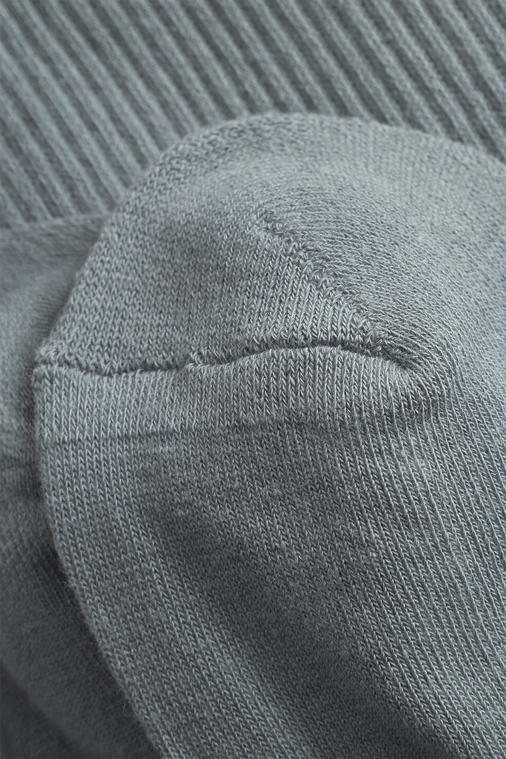 Close-up of the heel of a Gabba Zino Socks - Dust Sage Green cotton blend sock, featuring visible knit texture and stitching, resting on another matching ribbed fabric.