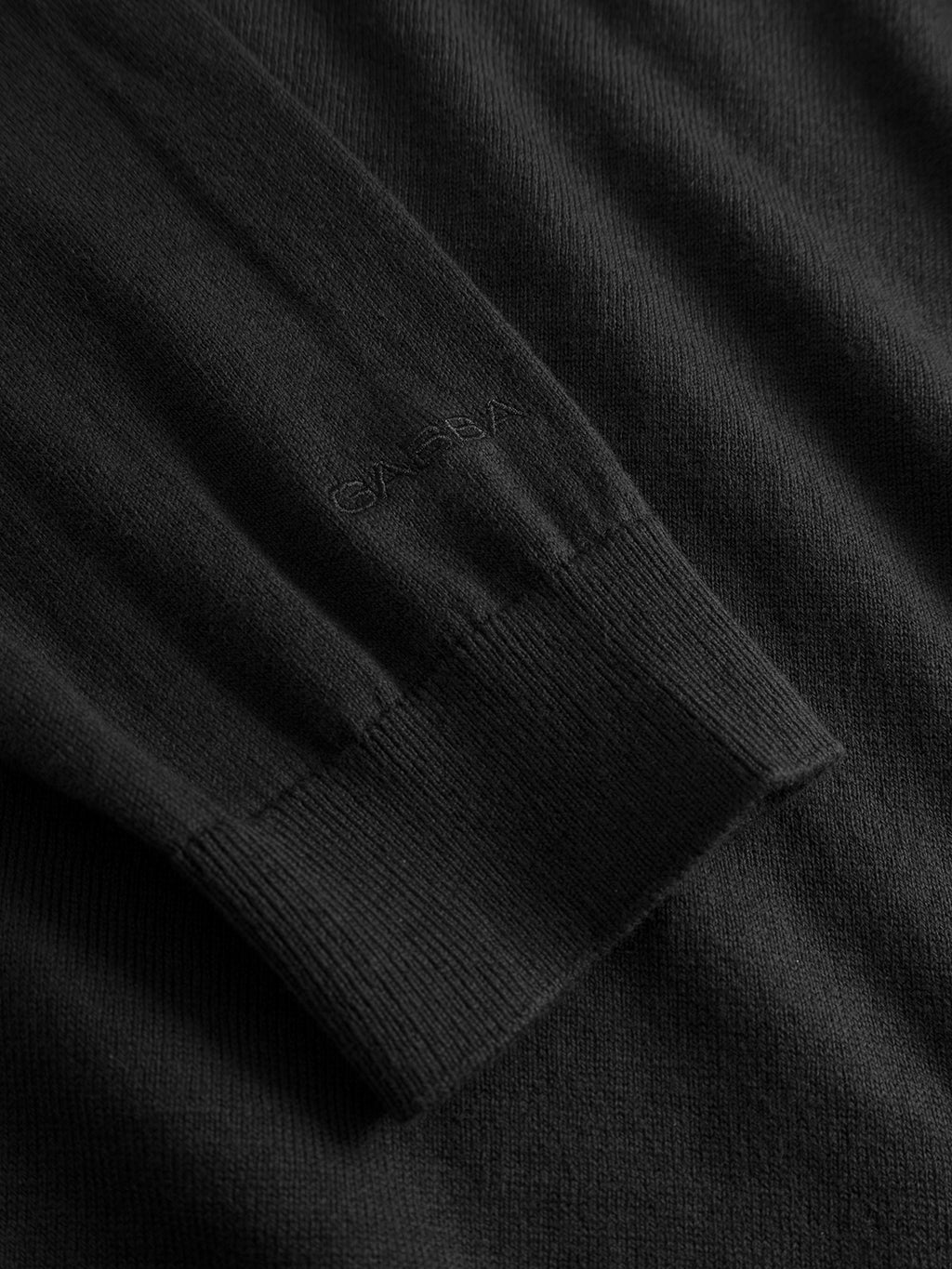 A close-up of the Aris Prex - Black sweater by Gabba shows its subtle ribbed cuff and matching black Gabba embroidery near the sleeve—perfect for elevating your classic roll neck look.