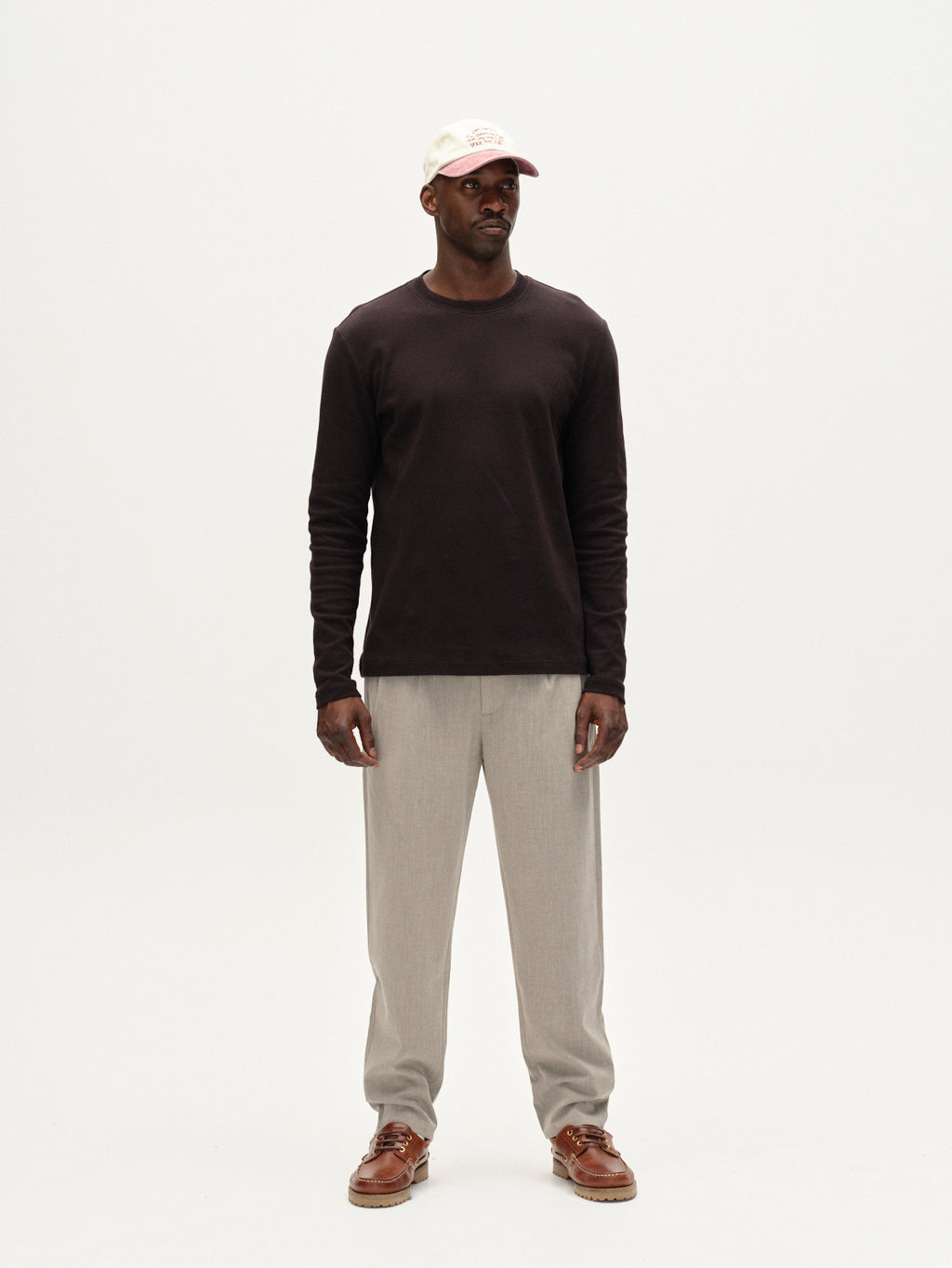 A man stands against a plain white background, facing forward with his hands by his sides. He wears Gabba Firenze Phin Cropped GRS trousers in sand, a long-sleeved dark shirt, a white cap, and brown shoes.
