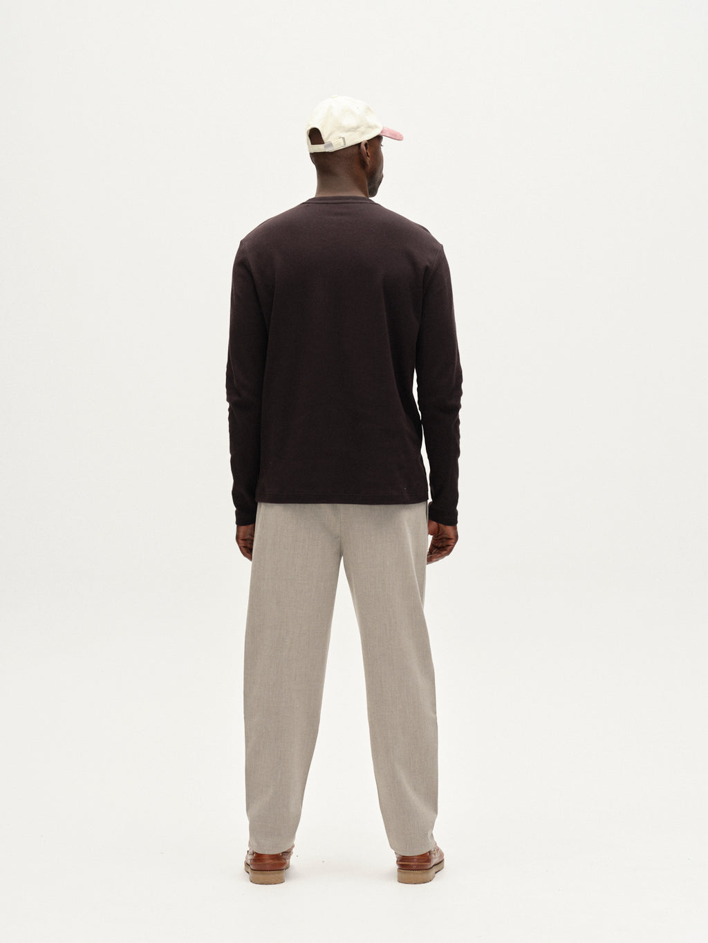 A man faces away from the camera wearing Gabbas Firenze Phin Cropped GRS trousers in Sand, styled with a white cap, pink brim, dark long-sleeved shirt, and brown shoes against a plain white background.