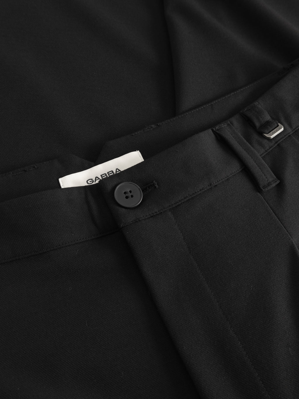 Close-up of GABfirenze Phin Cropped GRS - Black trousers by Gabba, featuring a black button, belt loop, and a white label—crafted for formal looks with a modern edge.