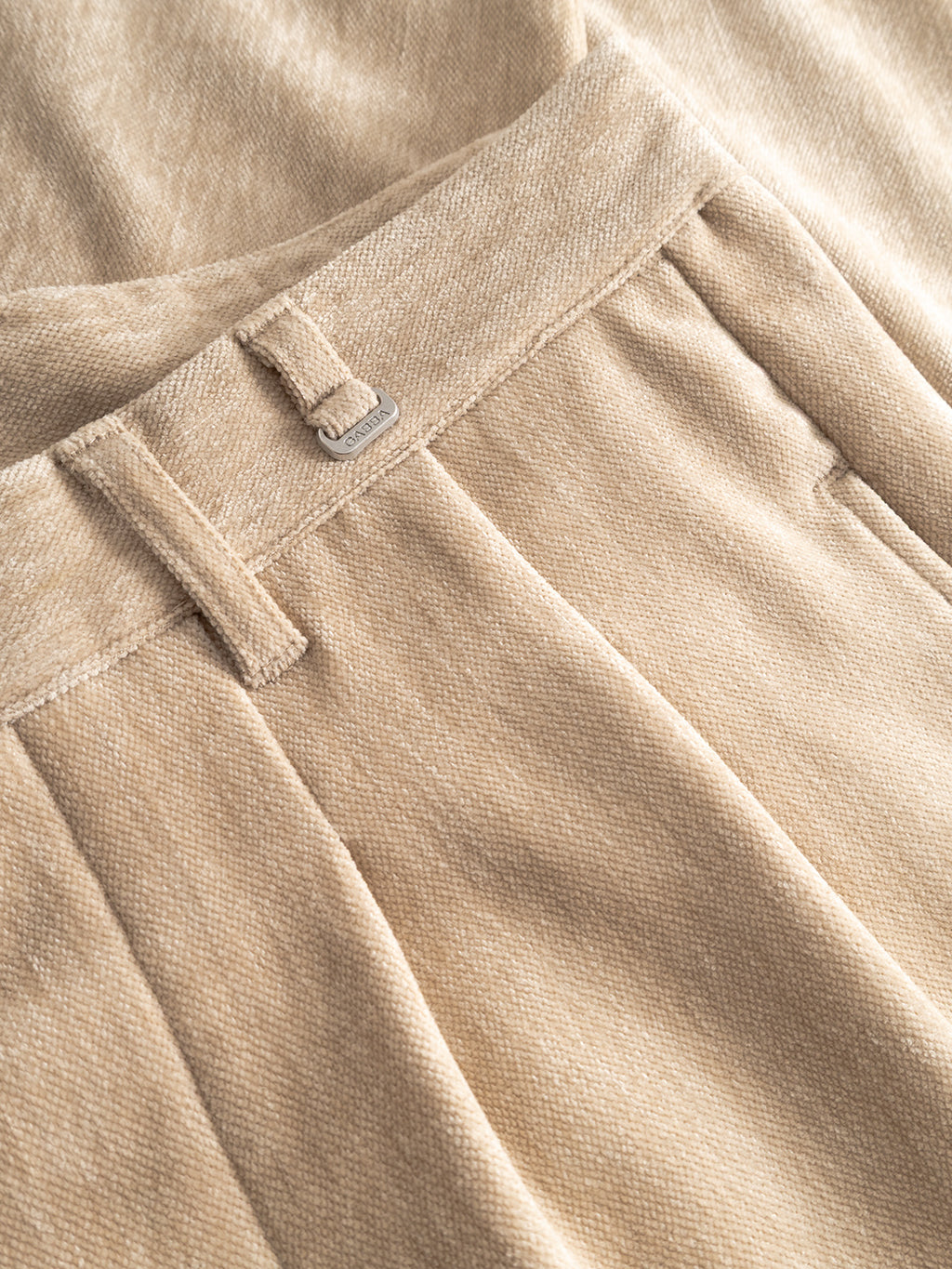 Close-up of Gabbas Firenze Velour pants in Lt. Sand, showcasing a luxurious feel with waistband, belt loop, metal tag, and pleats, styled on matching fabric.
