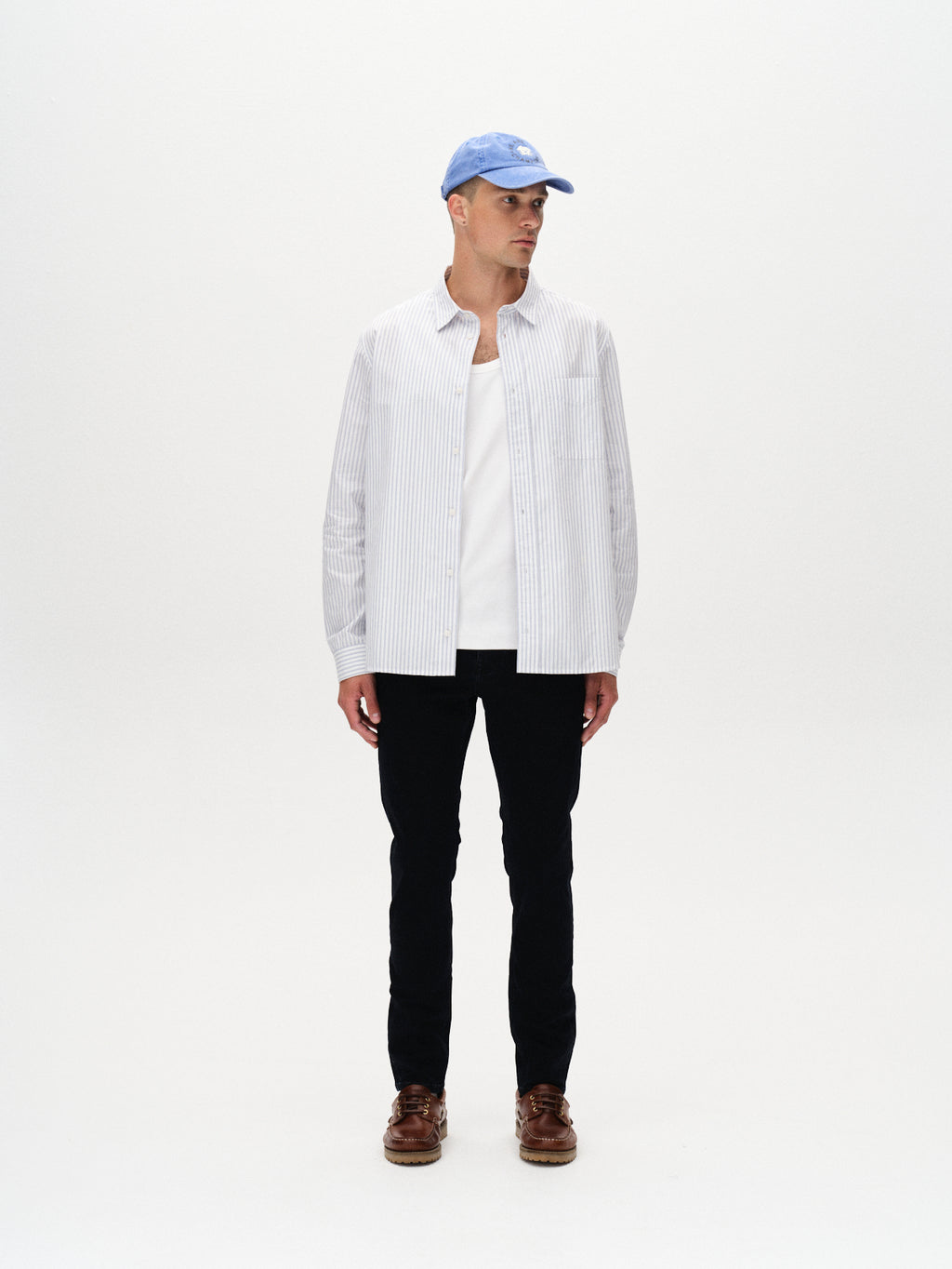 A man stands against a white background, wearing Gabbas Jones BlueBlack - Blue Black Denim slim straight fit jeans, a light blue cap, a white and gray striped button-up over a white t-shirt, and brown shoes. He looks slightly to his left.