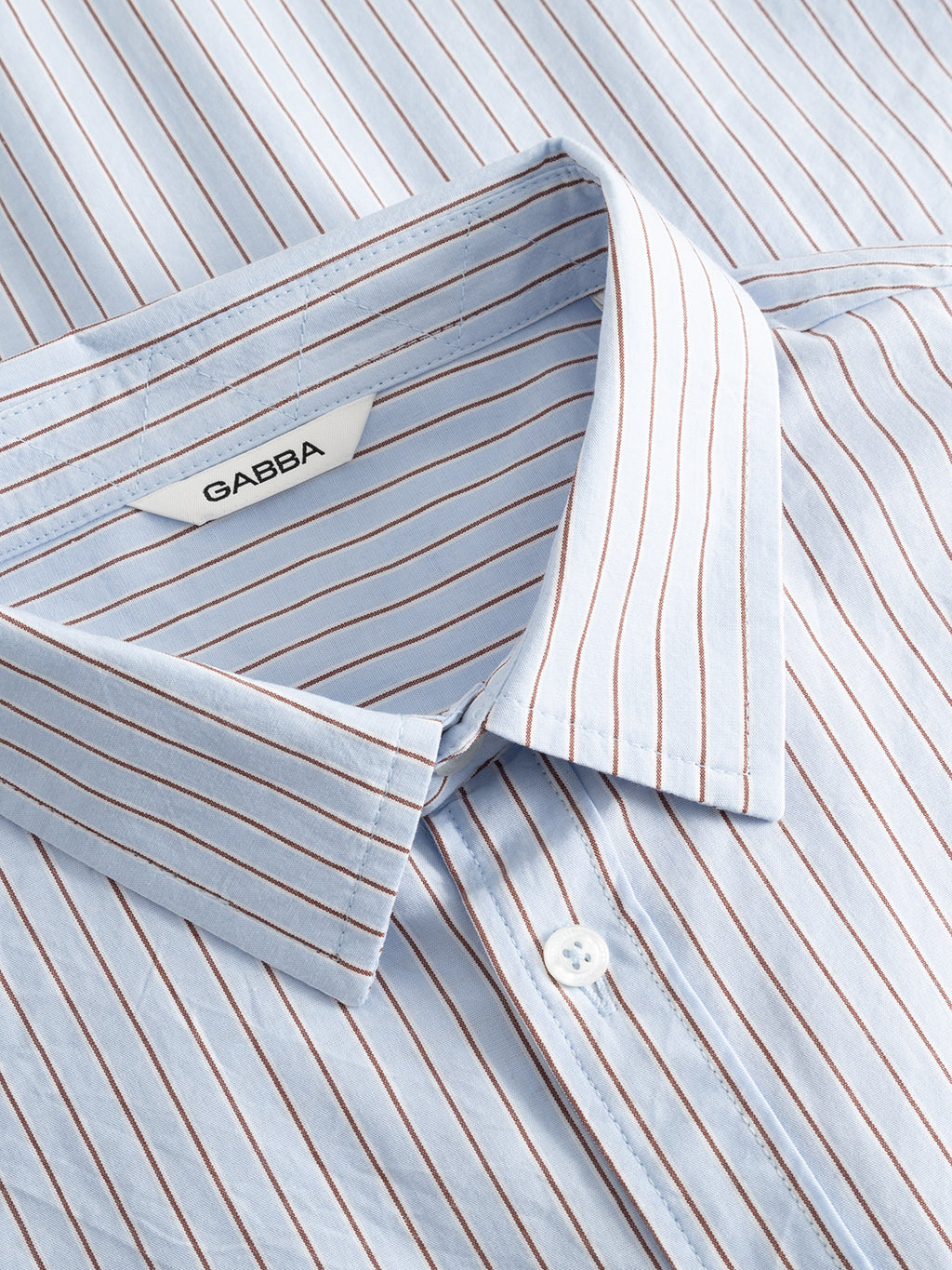 A close-up of the Gabba GABjens Over Bisi SS Shirt in Brown Stripe, a relaxed fit cotton shirt with light blue and thin brown vertical stripes, pointed collar, white button, and a Gabba label.