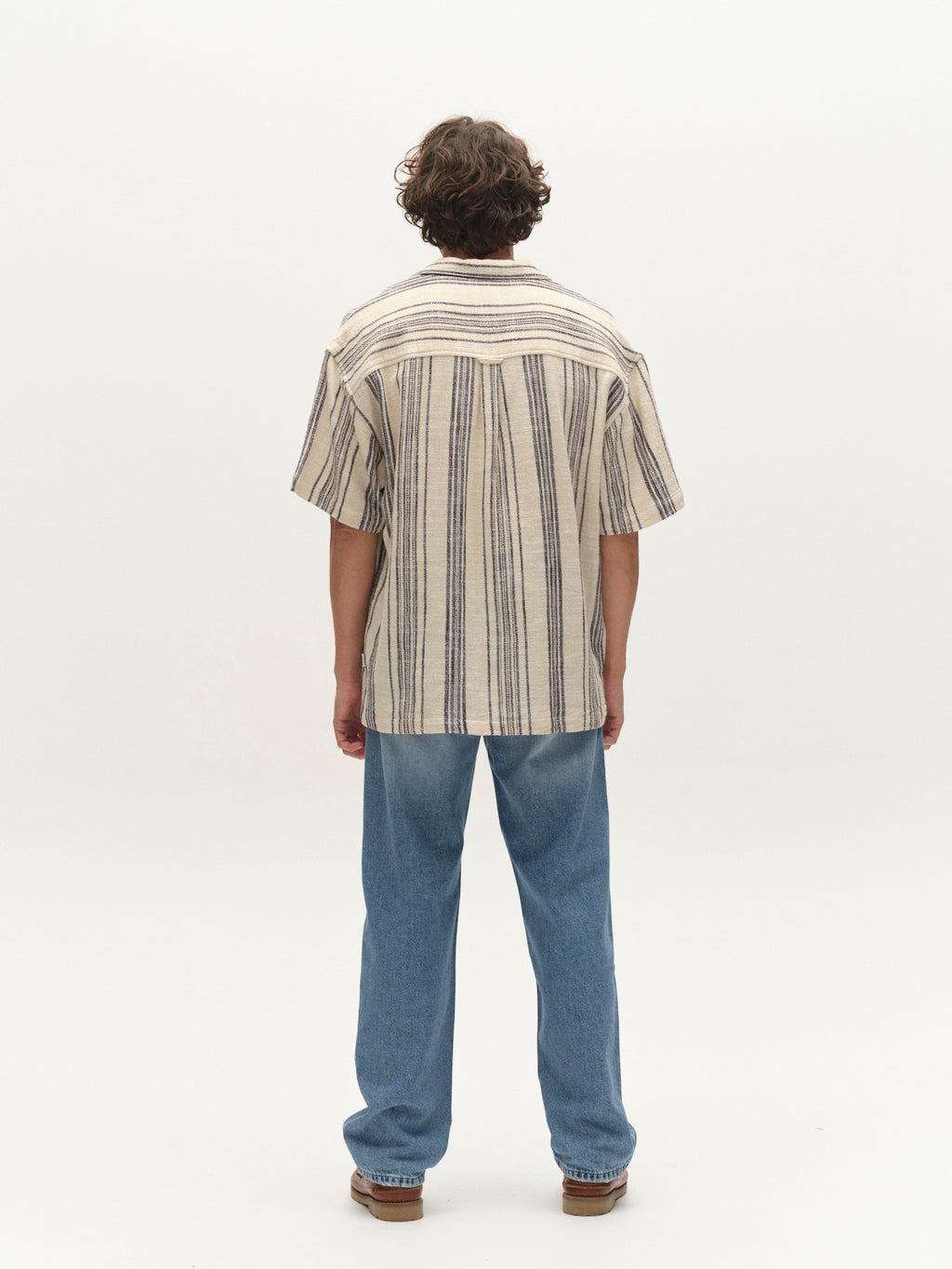 A person stands facing away from the camera on a plain white background, wearing the Gabba GABjens Over Resort Riga SS Shirt in Navy Stripe, paired with light blue jeans and brown shoes.