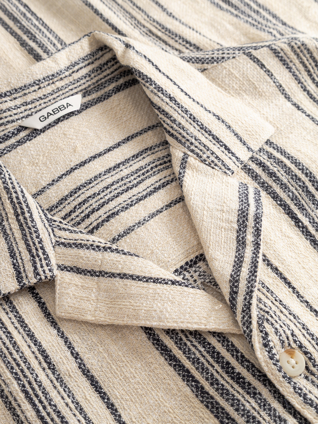 A close-up of the Gabba GABjens Over Resort Riga SS Shirt in navy stripe, featuring beige slub cotton fabric with navy blue stripes, a collar, a button, and a visible Gabba label.