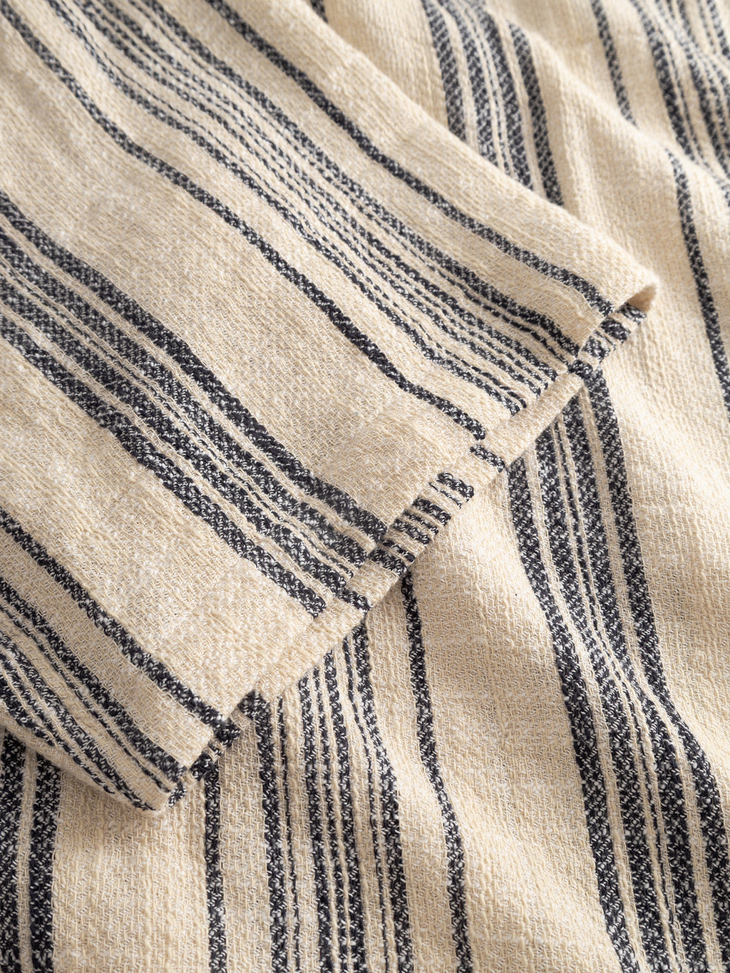 A close-up of the Gabba GABjens Over Resort Riga SS Shirt in Navy Stripe shows soft, beige woven fabric with dark blue vertical stripes and clean, unfinished edges, highlighting its textured, slub-cotton shirt design.