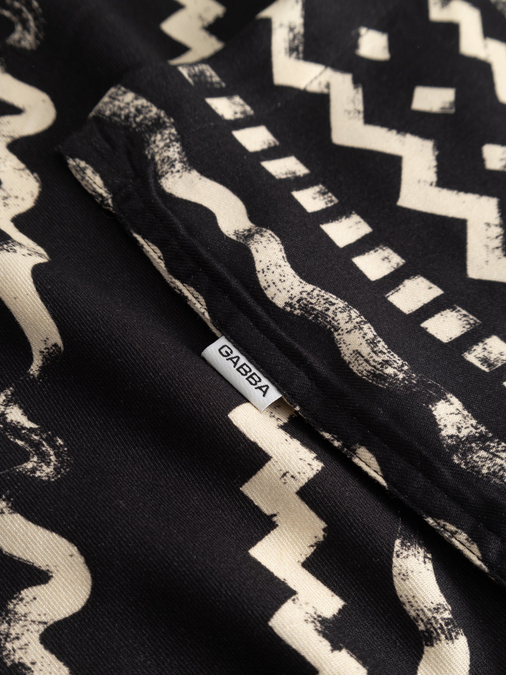 Close-up of the Gabba GABsven Reg Resort Oro SS Shirt in multi pattern, showing black viscose twill with off-white zigzag and dotted geometric prints. A small white Gabba tag is sewn into the seam.