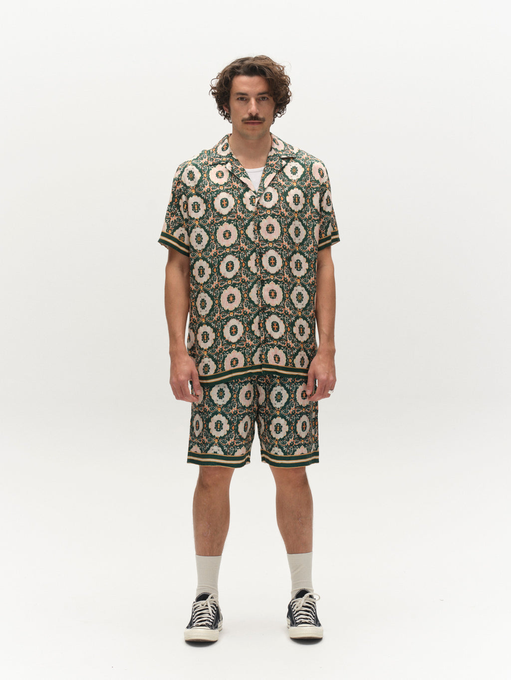 A man with curly hair and a mustache stands in a studio wearing the Gabba GABsven Reg Resort Oro SS Shirt in a green geometric pattern, paired with matching shorts, white socks, and black-and-white sneakers.