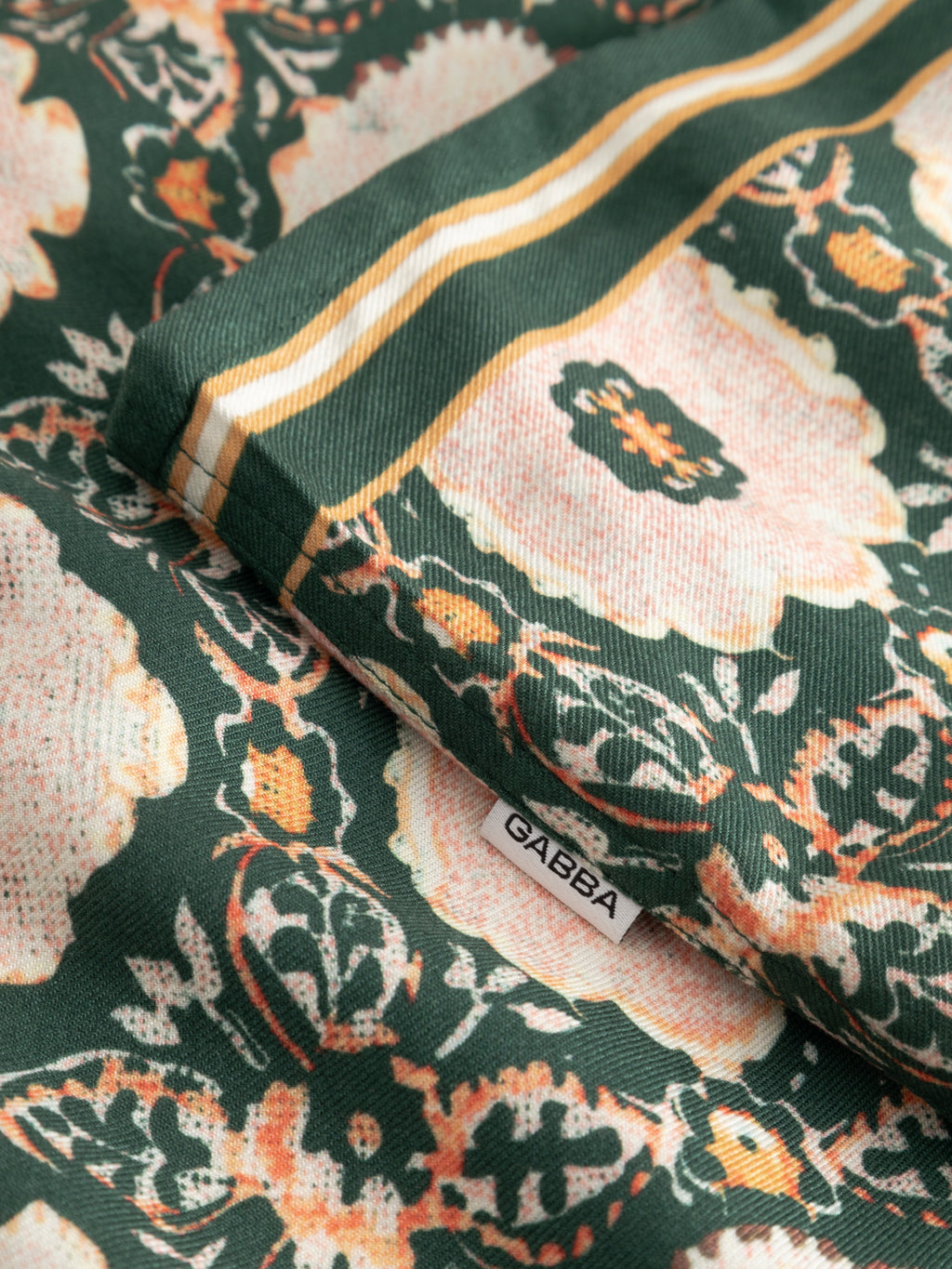 Close-up of the Gabba GABsven Reg Resort Oro SS Shirt - Green Pattern, featuring pink and orange floral designs, gold and white stripes, and a small white tag labeled 