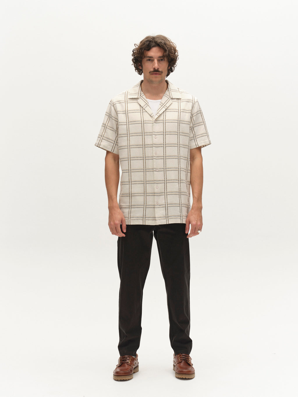 A man with curly hair and a mustache stands facing the camera, wearing the Gabba GABsven Reg Resort Wire SS Shirt in Beige Check, dark pants, and brown shoes against a plain white background.