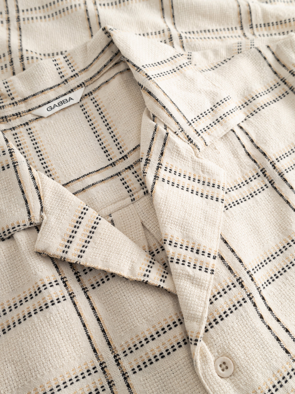 Close-up of the Gabba GABsven Reg Resort Wire SS Shirt in beige check, showing its textured black and tan plaid cotton, resort collar, and the visible Gabba label inside the neckline.