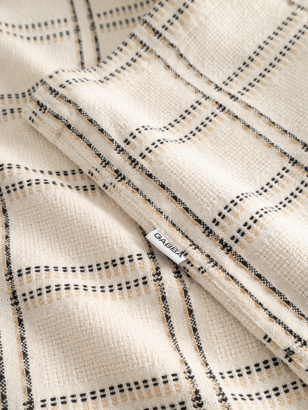 Close-up of the Gabba GABsven Reg Resort Wire SS Shirt in beige check, showing the cotton plaid fabric and a visible white Gabba tag sewn into the seam with detailed stitching.
