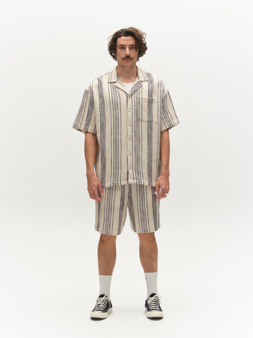 A man with curly hair and a mustache wears a short-sleeve, button-up shirt and Gabba GABtommi Riga Shorts in navy stripe, featuring vertical beige and navy stripes and an elasticated waistband. He stands facing forward in white socks and black sneakers.