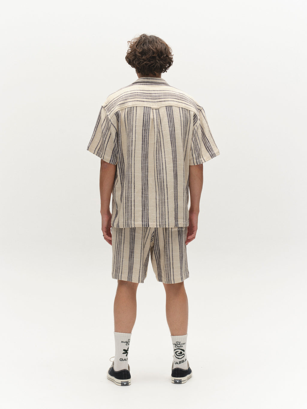 A person faces away, wearing Gabba’s GABtommi Riga Shorts in Navy Stripe with an elasticated waistband, a matching short-sleeve shirt, white socks with black text, and black sneakers, against a plain white background.
