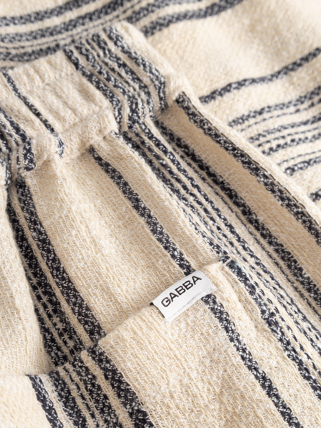 A close-up of GABtommi Riga Shorts - Navy Stripe by Gabba shows beige textured fabric with navy stripes and a pocket featuring a small white label with “GABBA” in black text.