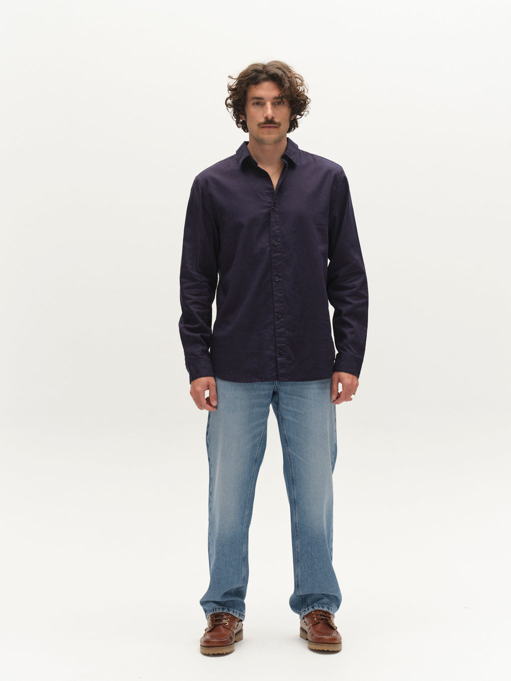 A man with curly hair and a mustache stands facing forward in a Gabba GABsven Reg Lins LS Shirt - Navy, paired with light blue jeans and brown shoes against a plain white background.