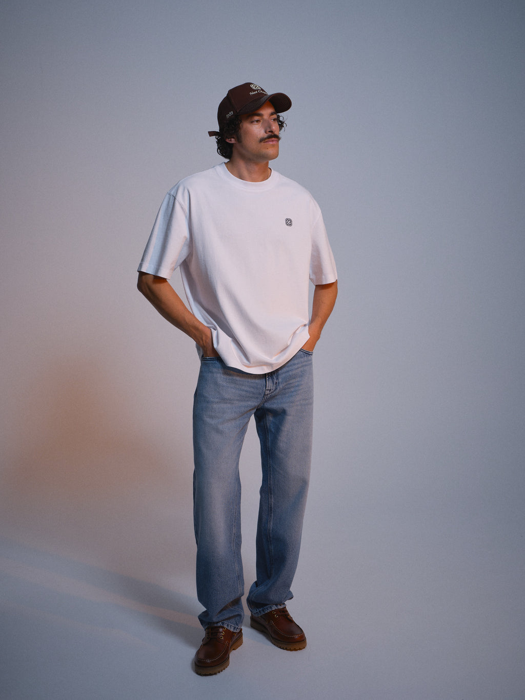 A man with a mustache stands against a plain background, wearing the Gabba GABlegin Wix Chap Boxy Hour SS T-Shirt in white, with a brown cap, light blue jeans, and brown shoes. He has his hands in his pockets and looks slightly to the side.