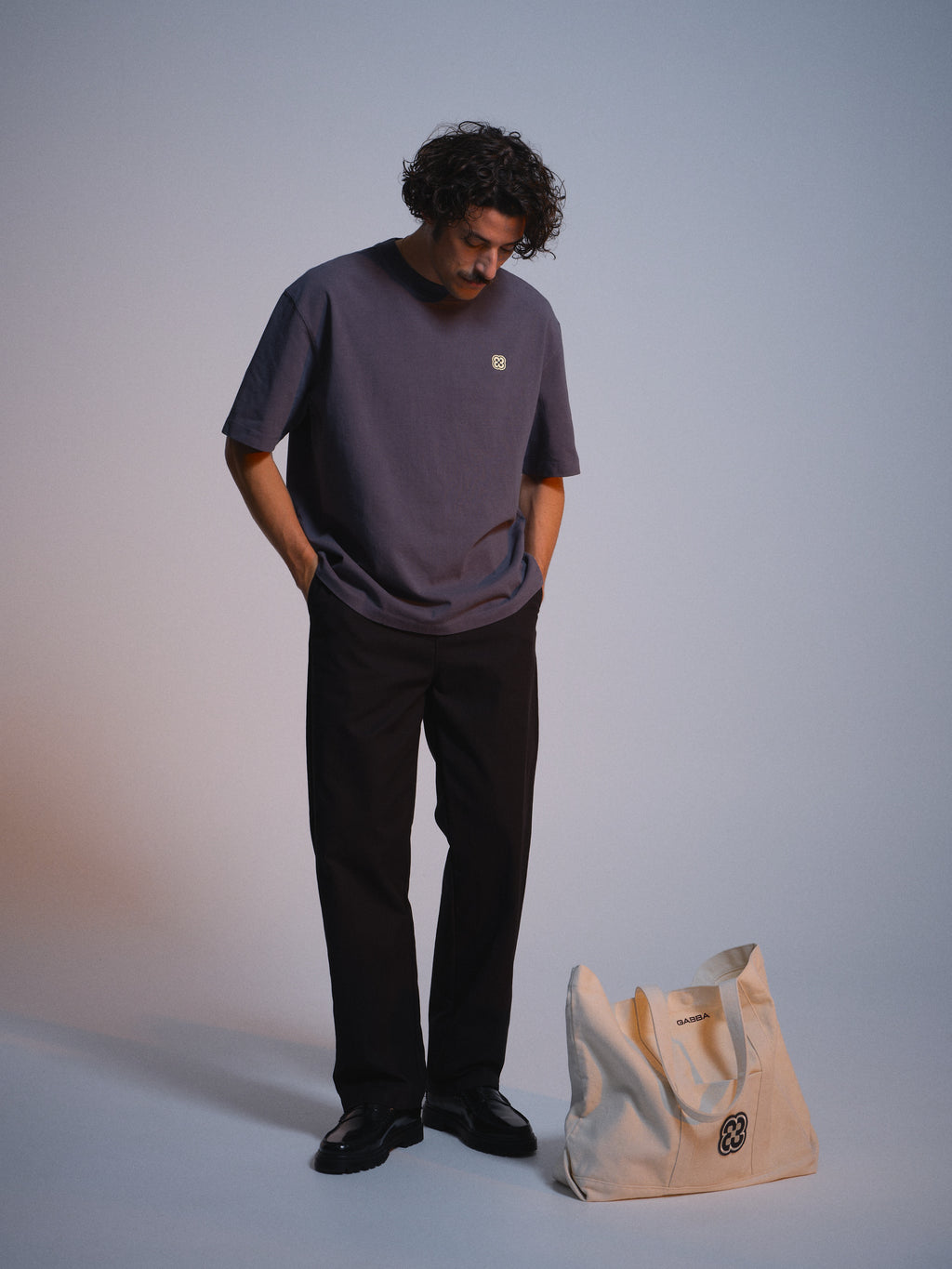 A man with curly hair wears a Gabba GABlegin Wix Chap Boxy Hour SS T-Shirt in Turbulence Blue, paired with black pants and shoes. He stands looking down with his hands in his pockets. A cream tote bag rests on the floor in front of him.