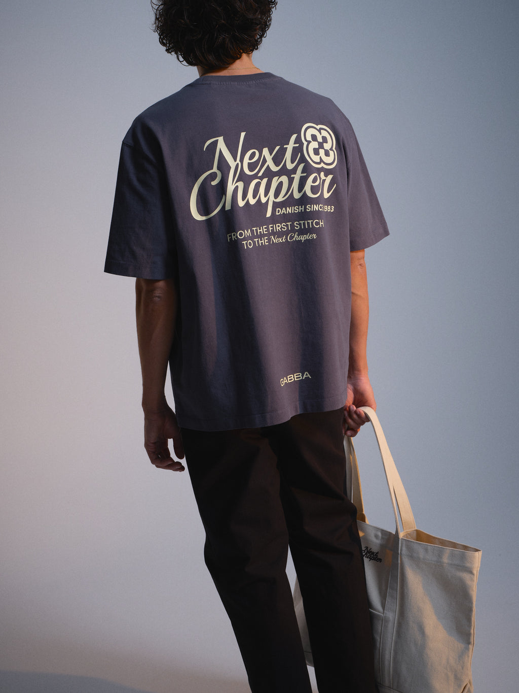 A person faces away, wearing the Gabba GABlegin Wix Chap Boxy Hour SS T-Shirt in Turbulence Blue with a bold back graphic and 