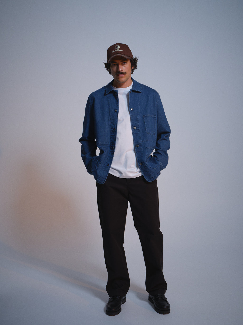 A person wears a brown cap, Gabba GABbeta Denim LS Overshirt in Mid Blue Denim with front pockets, a white t-shirt, black pants, and black shoes, standing with hands in pockets against a plain light background.