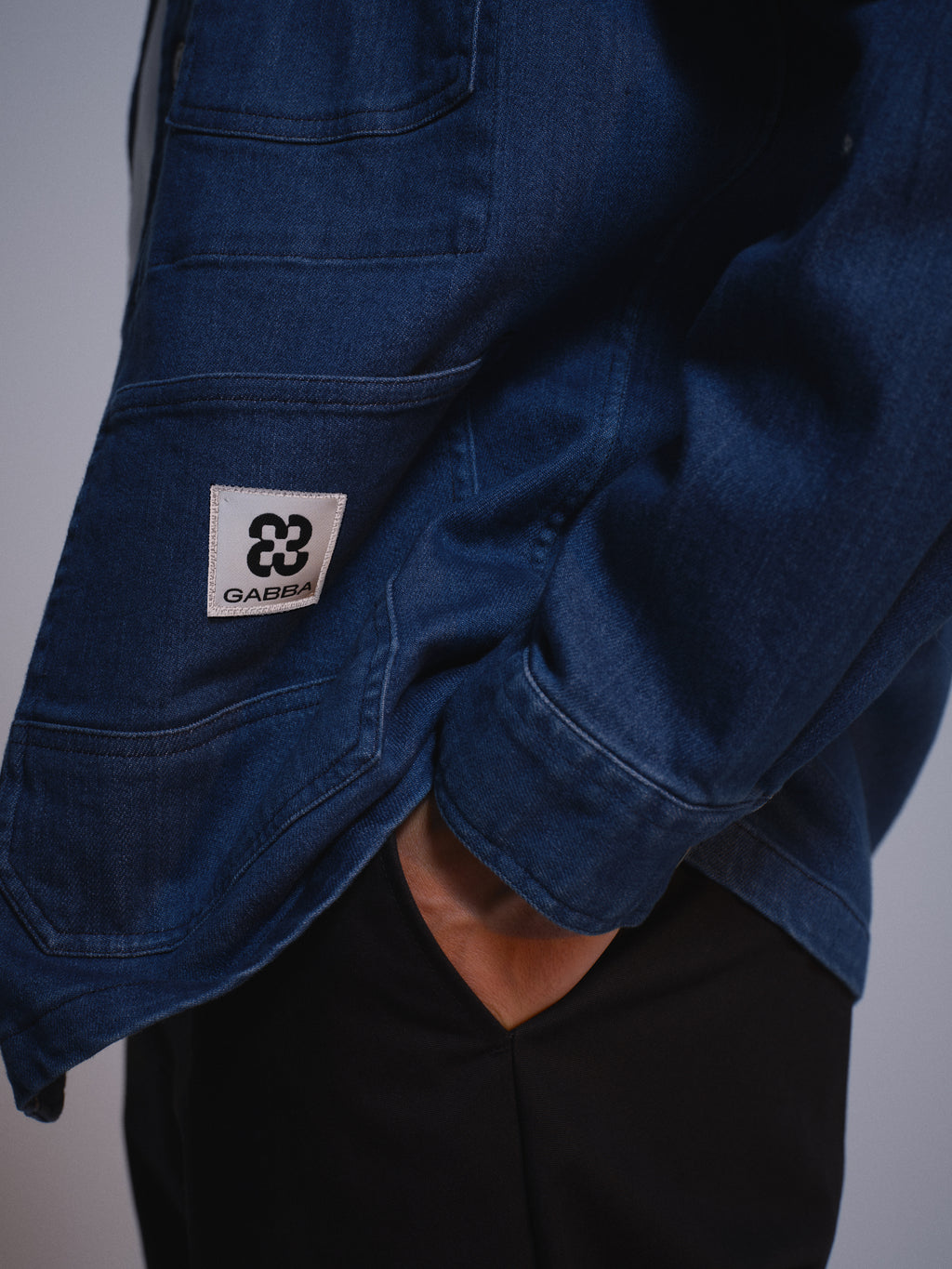 A person wearing the Gabba GABbeta Denim LS Overshirt in mid blue, featuring a white Gabba logo patch above the front pocket, paired with black pants and one hand casually in their pocket.