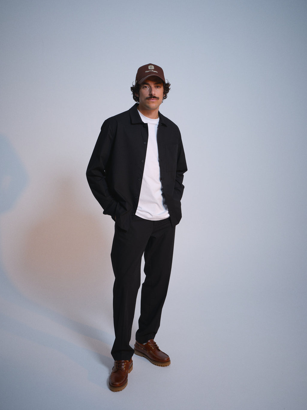 A mustached man in a brown cap, black jacket, white tee, Gabba GABjoe Tailor Dressy Pant in Black, and brown shoes stands with hands in pockets against a plain light background.