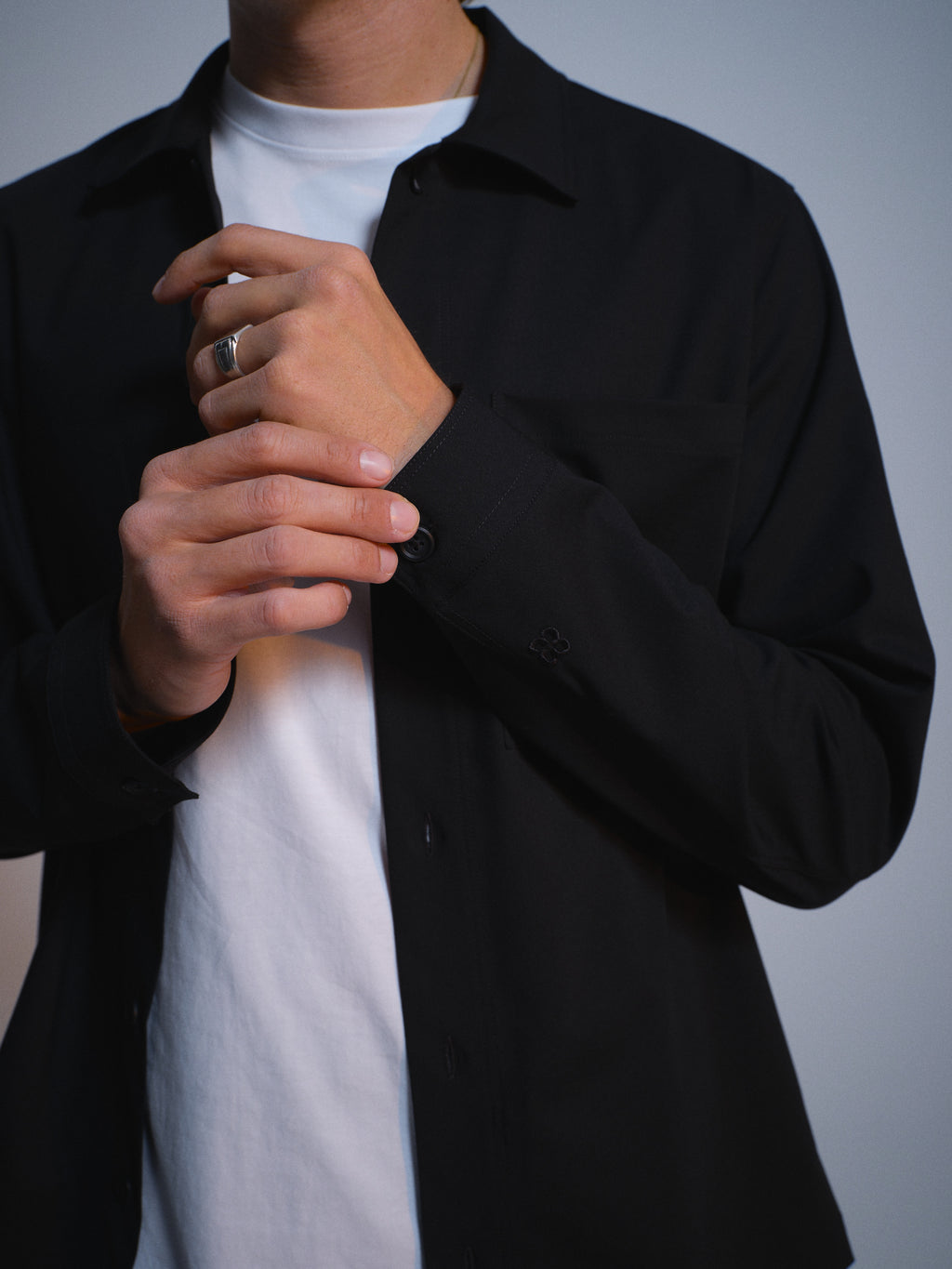A person wears the Gabba GABtogger Tailor Dressy Overshirt in black over a white t-shirt, adjusting their sleeve. The shot focuses on their upper body and hands, emphasizing smart layering and a silver ring on one finger.