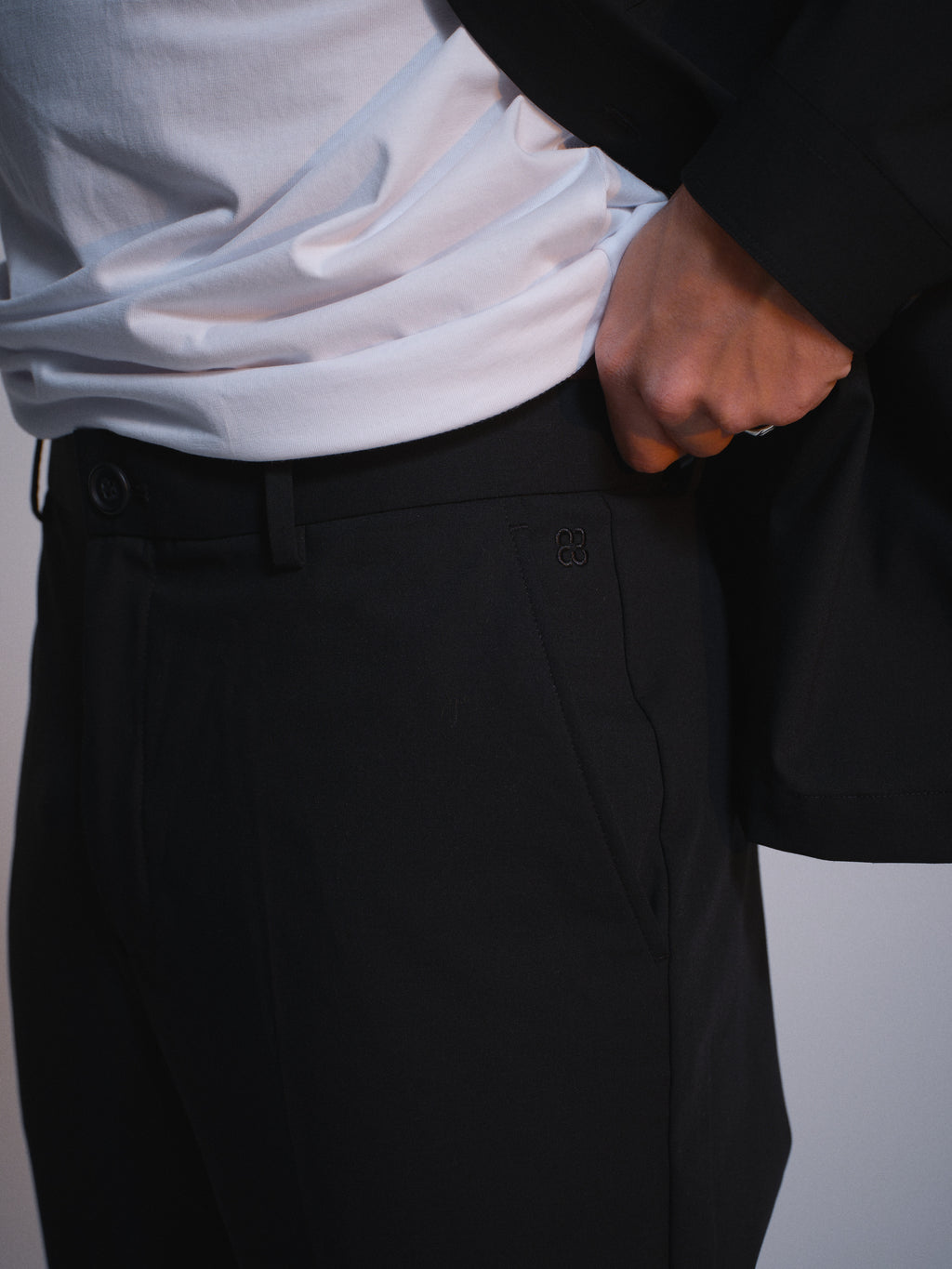 A person wears Gabba’s GABjoe Tailor Dressy Pant in black with a white t-shirt, hand in pocket and holding a black jacket. The shot highlights the waist and hips, set against a neutral background with subtle lighting.