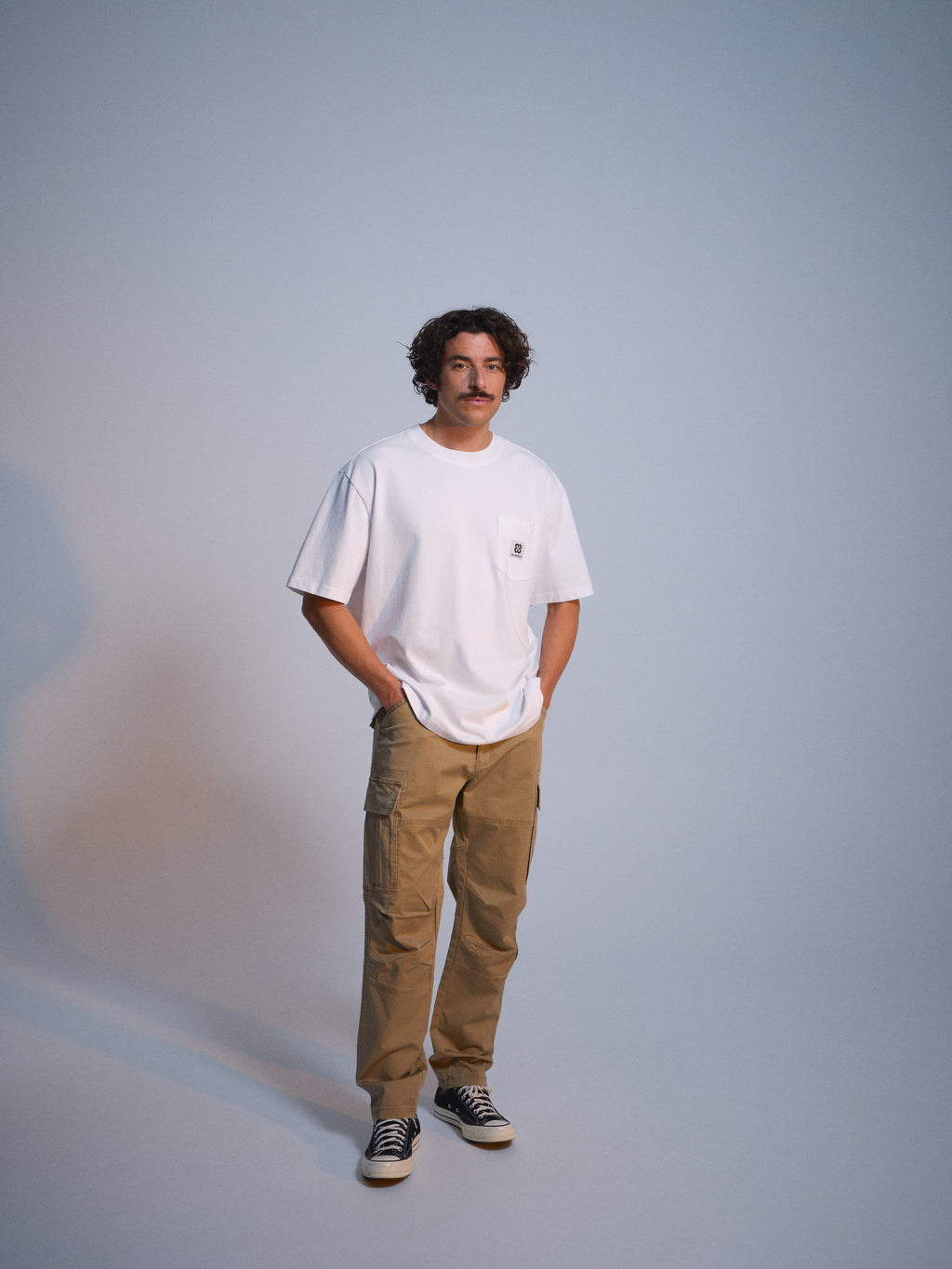 A man with curly brown hair and a mustache stands against a plain background, wearing a white t-shirt, Gabba GABjoe Vico Cargo Pant in Sand, and black-and-white sneakers, with his hands in his pockets.