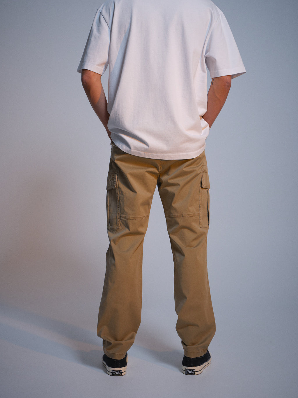 Someone wearing a white t-shirt, GABjoe Vico Cargo Pant - Sand by Gabba, and black sneakers stands facing away from the camera against a plain background.