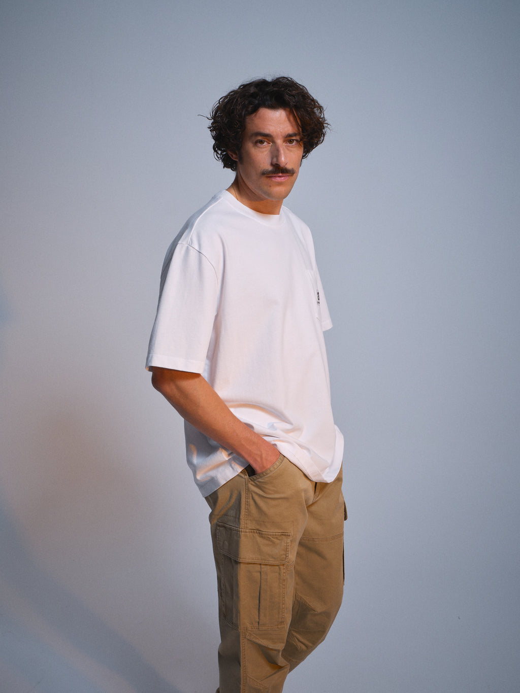 A man with curly hair and a mustache stands in a relaxed pose against a plain background, wearing a loose white t-shirt and Gabba GABjoe Vico Cargo Pant - Sand, with his hands in his pockets.