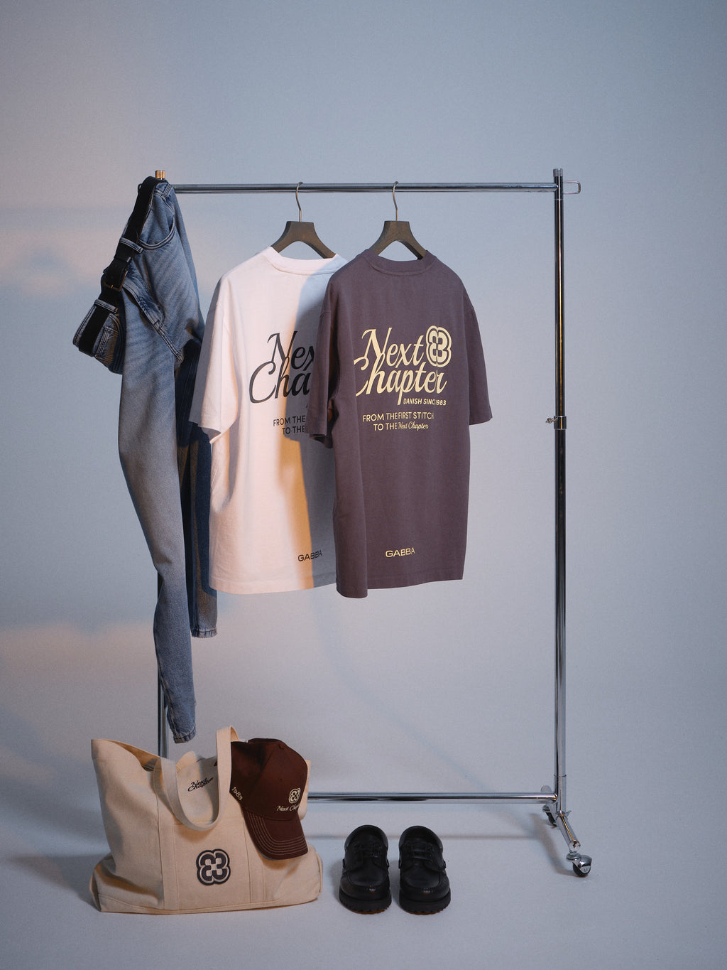 A clothing rack displays blue jeans, a white tee, and a gray “Next Chapter” shirt. On the floor is the GABchap Tote Bag - Off White by Gabba, paired with a brown cap and black sandals, all styled on a plain backdrop.