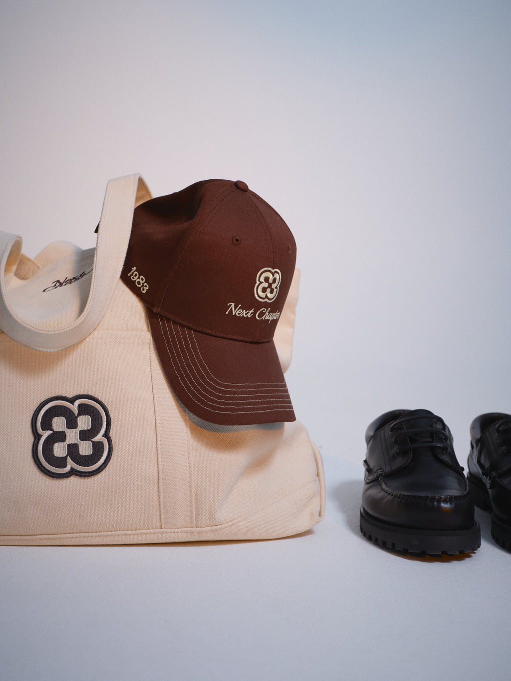 The Gabba GABchap Tote Bag in off white, a brown cap with yellow embroidery, and black leather shoes are displayed on a white surface.