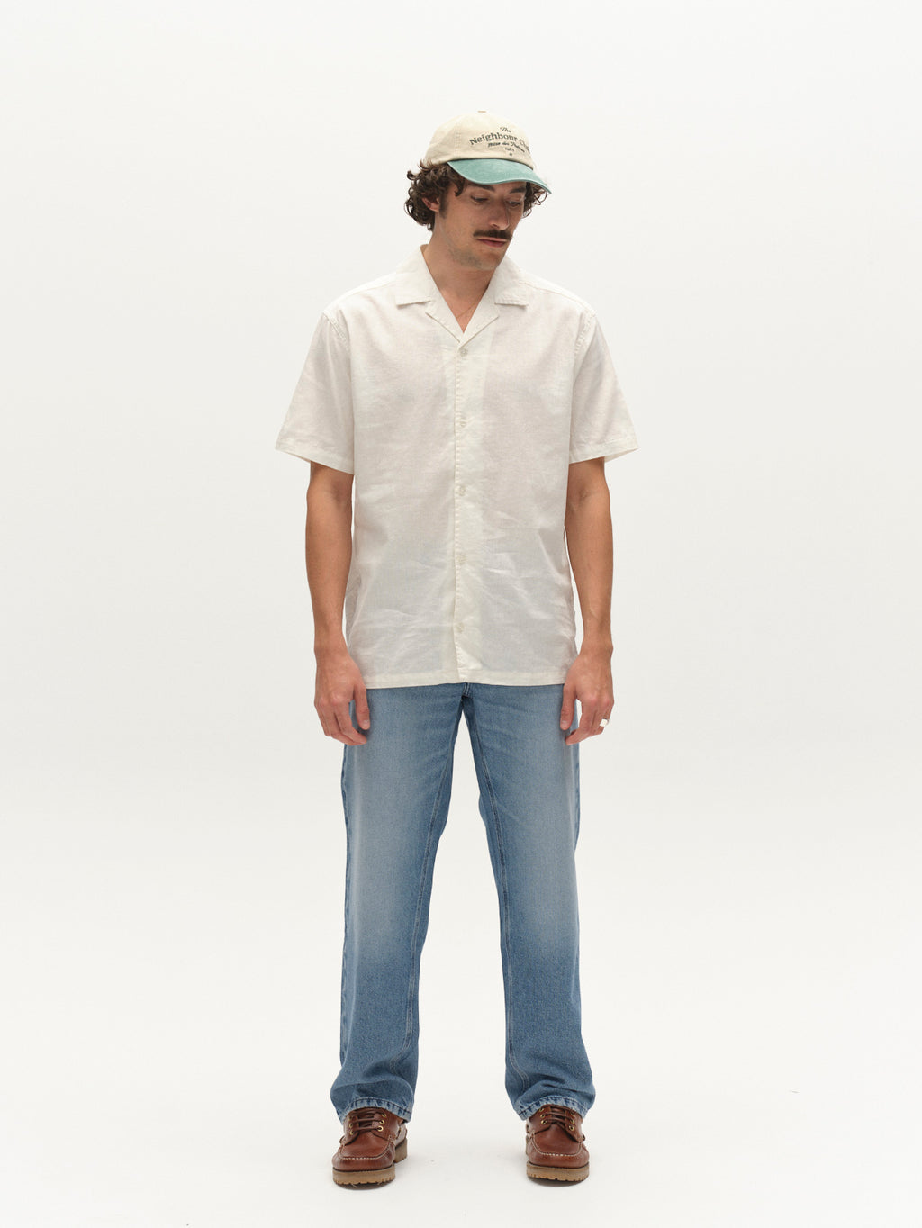Wearing the Gabba GABtimo Resort Lins SS Shirt in Off White, a person stands against a plain white background, paired with light blue jeans, brown shoes, and a beige cap, looking slightly downward with hands by their sides.